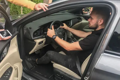 Mężczyzna siedzi za kierownicą, pokazując, jak ustawić fotel kierowcy. Dwie osoby pomagają mu, jedna trzyma telefon, druga opiera się o drzwi.