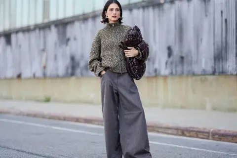 Stylisches Outfit: Eine Frau kombiniert eine graue Hose mit einem gemusterten Strickpullover und einer voluminösen Tasche.