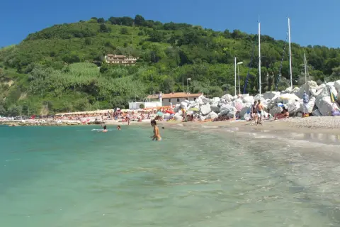 Spiaggia affollata con persone che nuotano e prendono il sole, sullo sfondo una collina verdeggiante. Gabicce Mare, dove si trova questo angolo di paradiso, è una perla dell'Adriatico.