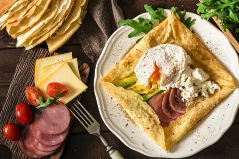 Pyszne crepes z jajkiem w koszulce, awokado, szynką i serem. Obok stos naleśników i dodatki.