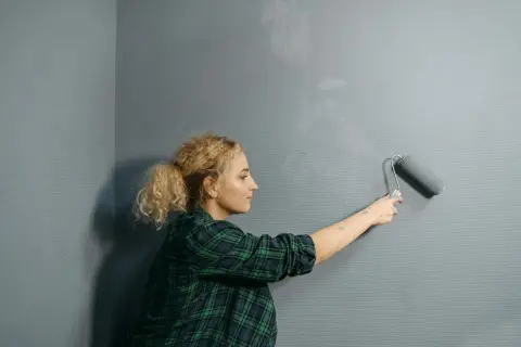 Mujer rubia con coleta pinta pared gris con rodillo. Se pregunta cuanto cuesta pintar un piso.