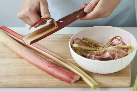 Hände schälen Rhabarberstangen auf einem Holzbrett. Die abgezogenen Streifen landen in einer Schüssel.