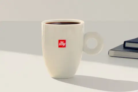Tazza bianca con logo Illy, piena di caffè americano, pronta per una pausa studio a casa.