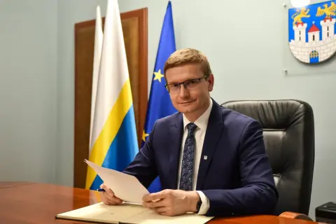 Mężczyzna w garniturze siedzi przy biurku, trzymając dokumenty. Za nim flagi i herb miasta, symbolizujące jego rolę w obszarze prawo administracyjne.