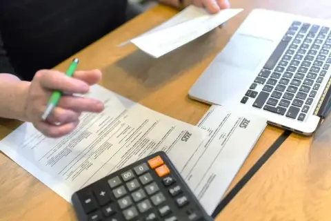 Dłoń z długopisem nad dokumentami ZUS, obok kalkulator i laptop.