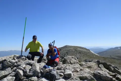 Dos excursionistas celebran en la cima tras una **cuerda larga ruta**. Un poste verde marca el camino.