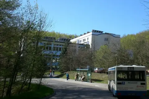 Sanatorium uzdrowiskowe „Leśnik” otoczone zielenią, z autobusem na pierwszym planie i ludźmi czekającymi na przystanku.