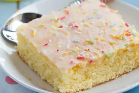 Ein Stück saftiger Fantakuchen vom Blech, mit Zuckerguss und bunten Streuseln bestreut, auf einem Teller mit Löffel.
