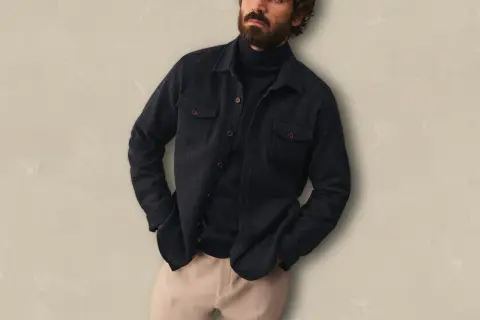 Un homme porte une surchemise foncée ouverte sur un col roulé bleu marine, avec un pantalon beige. Idéal pour savoir comment porter une surchemise avec style.