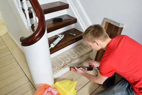 Man renoverar trappa med fogpistol och lim på trappsteg. Nya golvbrädor ligger redo.