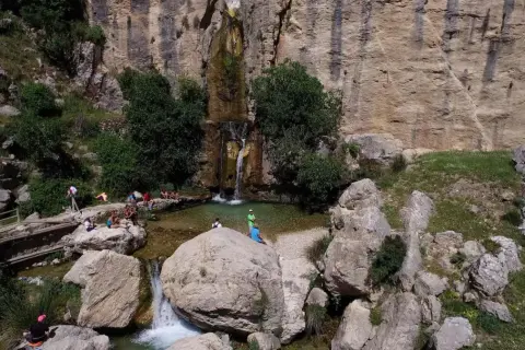 Un hermoso salto de agua cae en una poza natural rodeada de rocas y vegetación. Varias personas disfrutan del paisaje y del agua.