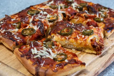 En saftig pan pizza med pepperoni och jalapeños, toppad med riven ost, ligger på en träskärbräda.