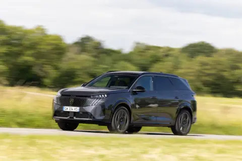 Ciemnoszary Peugeot 5008 pędzi po drodze, otoczony zielenią pól i drzew.