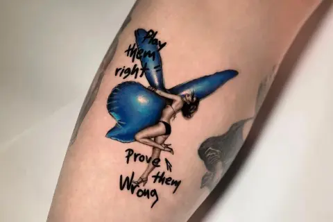 Ein Fineline Tattoo zeigt eine Frau mit Flügeln, die auf einem blauen Ballon sitzt. Darunter steht "Play them right, Prove them wrong".