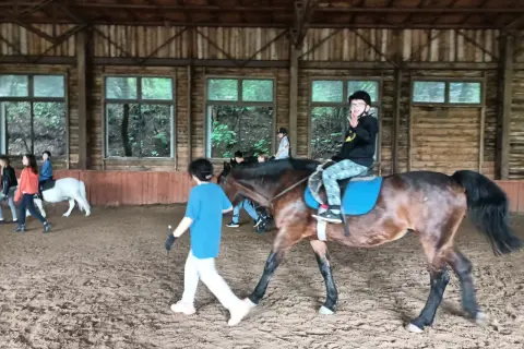 W stajni Black Horse Team Gdańsk, w owczarni, chłopiec na koniu macha do widza, a obok niego idzie instruktorka.