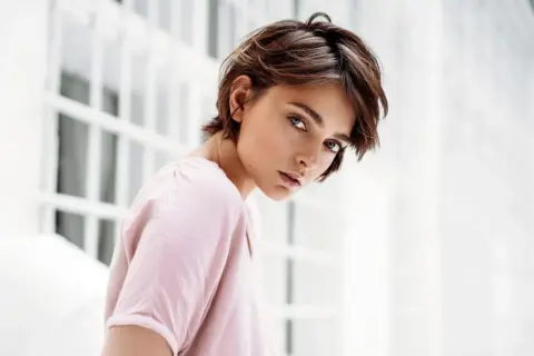 Junge Frau mit einem lässigen Pixie-Cut-Haarschnitt, blickt nachdenklich in die Kamera. Sie trägt ein zartrosa Oberteil.