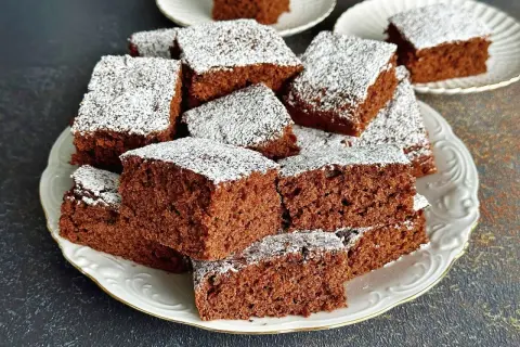 Pyszne ciasto czekoladowe pokrojone w kostkę, posypane cukrem pudrem, ułożone na białym talerzu.