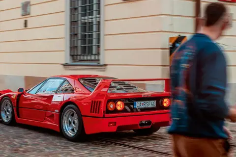 Czerwone Ferrari F40 z charakterystycznym spojlerem i okrągłymi światłami mija brukowaną ulicę, a obok przechodzi rozmyta postać mężczyzny.
