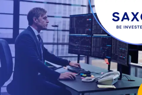 Mężczyzna w garniturze analizuje wykresy na monitorach w biurze Saxo Bank Polska. "Be invested" - hasło zachęca do inwestowania.