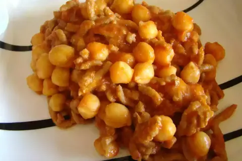 Un plato de garbanzos con carne picada, cocinado en una salsa rojiza y servido en una vajilla blanca con rayas negras.