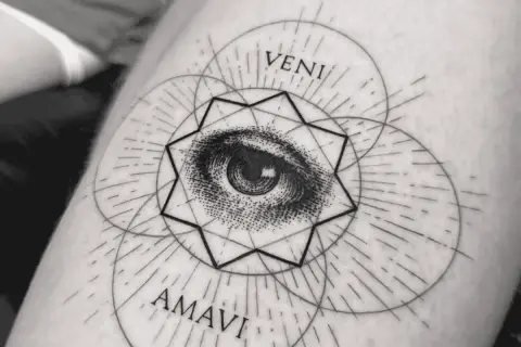 Ein filigranes Fineline Tattoo am Oberarm einer Frau zeigt ein Auge inmitten geometrischer Formen und der Worte "VENI AMAVI".