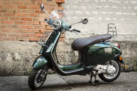 Elegancki, ciemnozielony skuter Piaggio z brązowym siedzeniem i chromowanymi dodatkami stoi na tle ceglanej ściany.