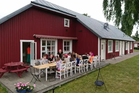 Gästerna njuter av en måltid utomhus vid Sjötorp vandrarhem. En grupp vänner samlas runt borden på altanen framför den röda byggnaden.