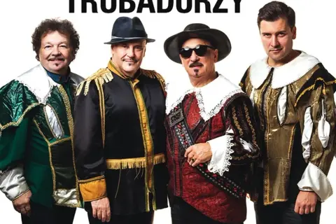 Czterech mężczyzn w barwnych strojach, przypominających stroje z epoki, pozuje do zdjęcia. Nad nimi widnieje napis "TRUBADURZY".