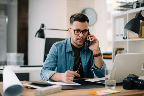 Mężczyzna w okularach rozmawia przez telefon przy biurku z laptopem i aparatem.