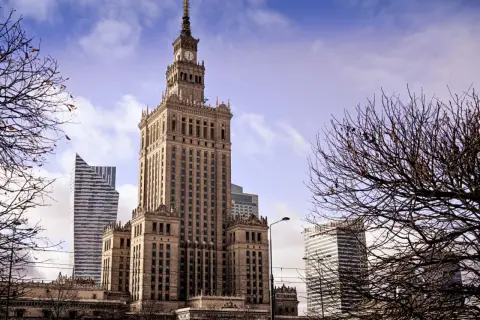 Kulturpalatset Warszawa reser sig stolt mot en molnig himmel, omgiven av moderna skyskrapor och kala trädgrenar.