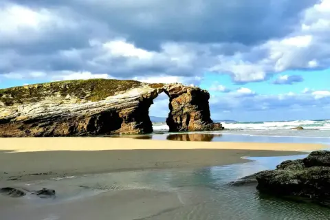 Arco natural en las playas de Galicia, con arena dorada, mar azul y cielo nublado.