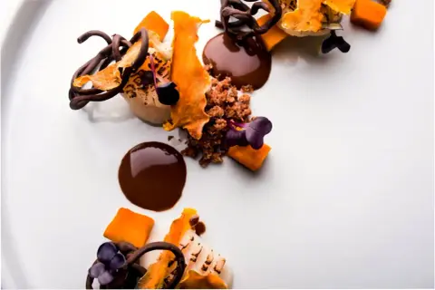 Postre de autor con chocolate, naranja y flores moradas. Una obra de arte culinaria que promete una experiencia gastronómica inolvidable.