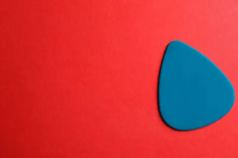 Ein blaues Plektrum liegt auf rotem Untergrund. Was ist ein Plektrum? Ein kleines Werkzeug zum Zupfen von Saiten.