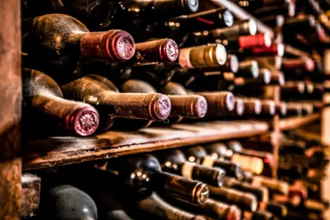 Des bouteilles de vin anciennes, poussiéreuses, sur des étagères en bois. Certaines ont des capsules rouges, d'autres dorées. On imagine le goût d'un vin qui pique en bouche.