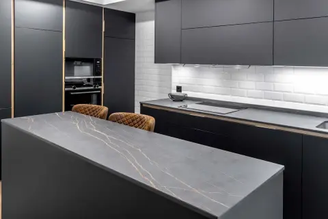 Isla de cocina moderna con encimera de piedra gris veteada en oro. Dos taburetes de cuero marrón esperan junto a la medida encimera.