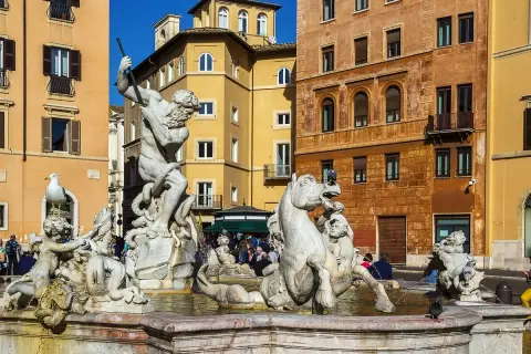 La Fuente de Neptuno en Roma, con su imponente estatua de mármol luchando contra un pulpo, rodeada de caballos marinos y figuras mitológicas, bajo el sol y edificios coloridos.