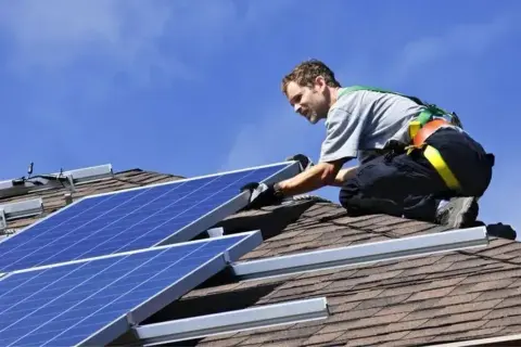 Installatore sul tetto, con imbracatura, che fissa pannelli fotovoltaici. L'orientamento dei pannelli è cruciale per massimizzare l'energia solare.
