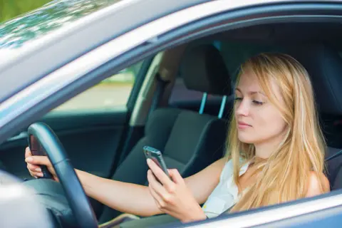 Mujer rubia al volante, mirando su móvil. Al distraerse el conductor tarda, poniendo en riesgo su seguridad y la de otros.