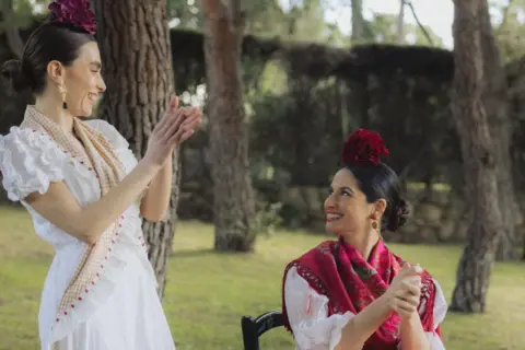 Dos mujeres vestidas para ir al Rocío, con trajes blancos y mantones, sonríen y aplauden en un jardín.
