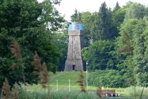 Kamienna wieża Bismarcka w Szczecinku, otoczona zielenią, z błękitnym elementem na szczycie.