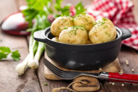 Młode ziemniaki w czarnej misce, posypane koperkiem, obok widelca i dymki.
