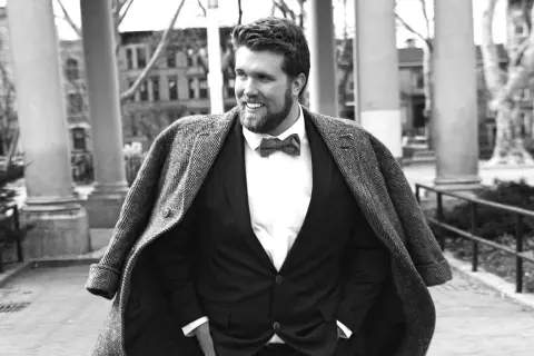 Un homme gros bien habillé, souriant, porte un costume sombre, une chemise blanche et un nœud papillon. Son manteau est jeté sur ses épaules.