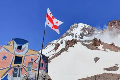 Kolorowy budynek i gruzińska flaga na tle ośnieżonych szczytów. Kazbek wysokość robi wrażenie.
