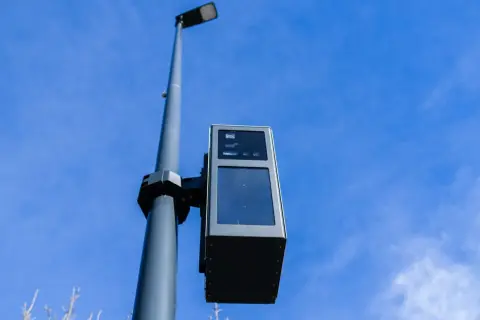 Un radar de ceinture, fixé sur un poteau, surveille la route sous un ciel bleu.