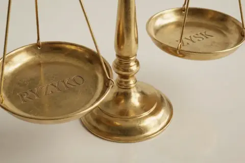 Złota waga szalkowa z napisami "RYZYKO" i "ZYSK". Emitent kto to? To pytanie o równowagę między ryzykiem a zyskiem.