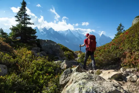 Randonneur avec sac à dos rouge, bâtons de marche, sur un sentier rocheux. Montagnes enneigées en arrière-plan. Combien de jours de marche pour atteindre ce sommet ? gr5.