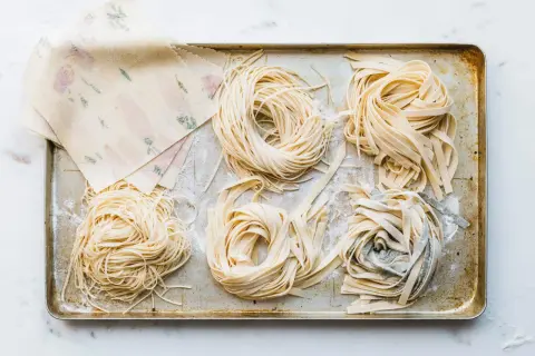 Frische Nudeln in verschiedenen Formen liegen auf einem Blech. Ein Stapel mit Blumenmuster verzierter Teigblätter liegt daneben. Perfekt für alle, die Nudeln selber machen wollen.