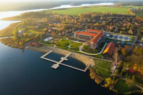 Widok z lotu ptaka na kompleks Lake Hill Mazury z jeziorem, plażą, molo i budynkami w jesiennej scenerii.