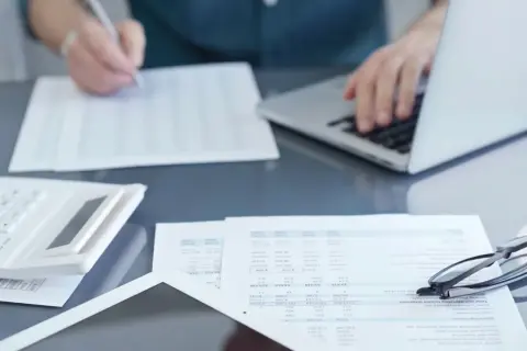 Osoba pracuje przy laptopie, przeglądając dokumenty i pisząc. Na biurku leży kalkulator i okulary. Przygotowania do podpisania umowy zlecenie 2026.