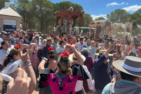Multitud celebra con alegría, bailando y cantando al paso de una carreta adornada. Se percibe el espíritu de una sevillana rociera en pleno fervor.